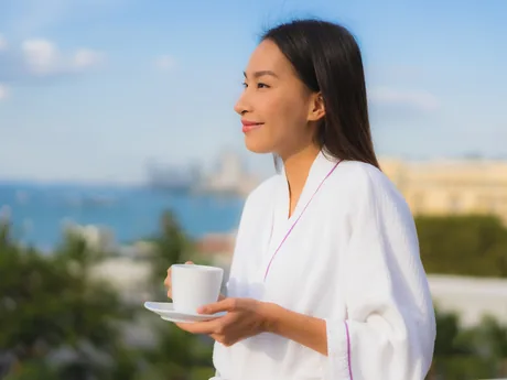 A woman in a robe holding a mug, looking out at the water