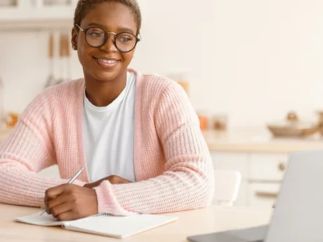 A woman at her kitchen table with a laptop, writing in a notebook