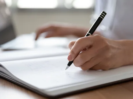 A close view of hands writing in a notebook beside a laptop