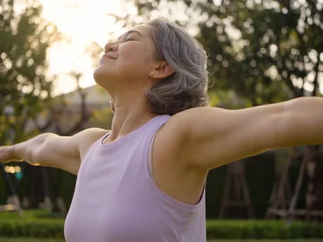 A woman with gray hair outdoors at sunset, arms open and eyes closed